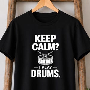 May include: A black short-sleeved t-shirt featuring white text that reads KEEP CALM? I PLAY DRUMS. The design includes a graphic illustration of a snare drum with two crossed drumsticks positioned directly above the drum in the centre.