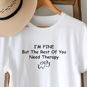May include: A white short-sleeved t-shirt featuring the black printed text I'M FINE But The Rest Of You Need Therapy and a small swirling graphic below the words. A tan fedora hat with a black band is positioned above the t-shirt on a hanger.