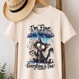 May include: A cream-coloured short-sleeve t-shirt featuring a graphic of a dishevelled black cat sitting in a puddle under a rain cloud with lightning. The cat holds a coffee mug, and the text reads I'm Fine and Everything is Fine.