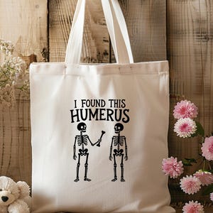 May include: A cream-coloured canvas tote bag featuring a black graphic print of two skeletons. One skeleton holds a bone towards the other, with the text "I FOUND THIS HUMERUS" printed in black lettering above the skeletons on the front of the bag.