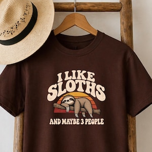 May include: A dark brown short-sleeve t-shirt featuring a graphic of a sloth resting on a branch against a sunset background. The text reads I LIKE SLOTHS AND MAYBE 3 PEOPLE. A tan straw hat with a black band is positioned above the t-shirt.