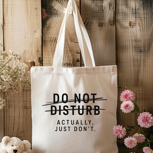 May include: A cream-coloured canvas tote bag featuring black text that reads DO NOT DISTURB with a line through it, followed by the text ACTUALLY, JUST DON'T. The bag has two long shoulder straps and is constructed from a plain, light-coloured fabric.