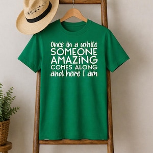 May include: A green t-shirt with the text "Once in a while SOMEONE AMAZING COMES ALONG and here I am" in white. A straw hat with a black band hangs above the shirt. The shirt is displayed on a wooden ladder.