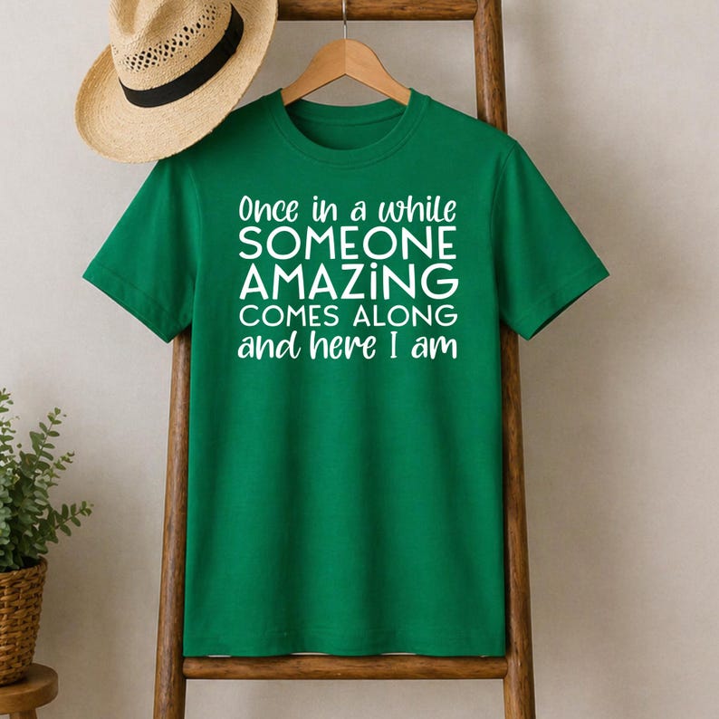 May include: A green t-shirt with the text "Once in a while SOMEONE AMAZING COMES ALONG and here I am" in white. A straw hat with a black band hangs above the shirt. The shirt is displayed on a wooden ladder.