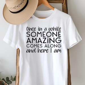 May include: White t-shirt with the text "Once in a while SOMEONE AMAZING COMES ALONG and here I am" in black. A tan hat with a black band hangs above the shirt on a wooden hanger.