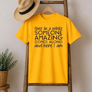 May include: Mustard yellow t-shirt with the text "Once in a while SOMEONE AMAZING COMES ALONG and here I am" in black. The shirt is hanging on a wooden hanger.