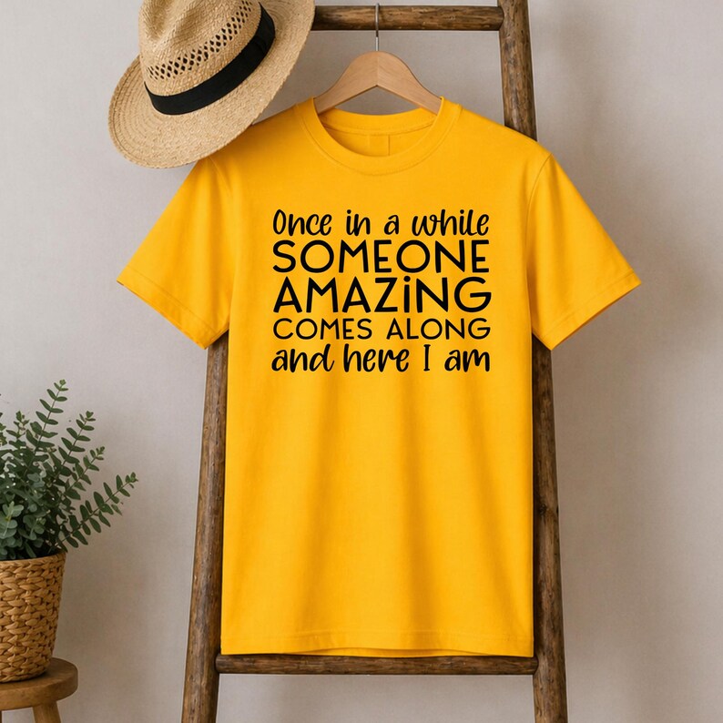 May include: Mustard yellow t-shirt with the text "Once in a while SOMEONE AMAZING COMES ALONG and here I am" in black. The shirt is hanging on a wooden hanger.