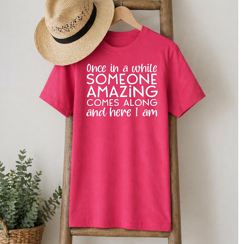 May include: A bright pink t-shirt with the text "Once in a while SOMEONE AMAZING COMES ALONG and here I am" in white. A straw hat with a black band hangs above the shirt.