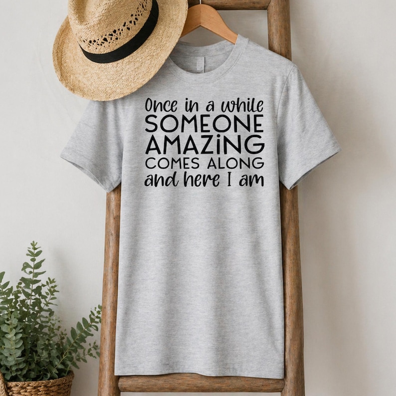 May include: A light gray t-shirt with the text "Once in a while SOMEONE AMAZING COMES ALONG and here I am" in black. A straw hat with a black band hangs above the shirt.