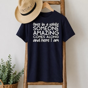 May include: Navy blue t-shirt with the text "Once in a while SOMEONE AMAZING COMES ALONG and here I am" in white. A straw hat hangs above the shirt on a wooden ladder.
