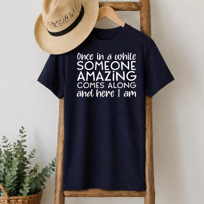 May include: Navy blue t-shirt with the text "Once in a while SOMEONE AMAZING COMES ALONG and here I am" in white. A straw hat hangs above the shirt on a wooden ladder.