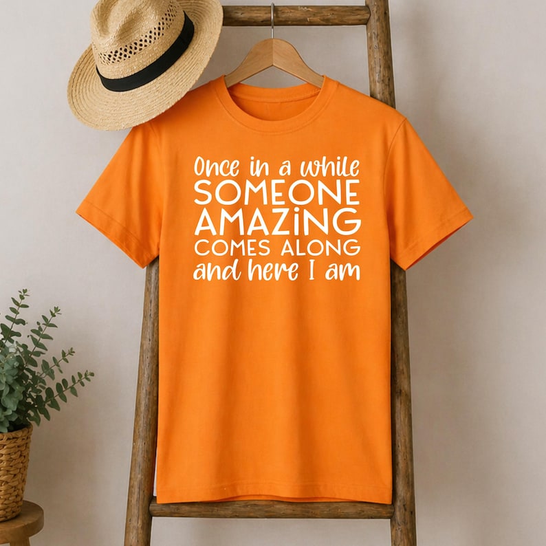 May include: Orange t-shirt with the text "Once in a while SOMEONE AMAZING COMES ALONG and here I am" in white. A straw hat with a black band rests on top of the shirt. The shirt is hanging on a wooden ladder.