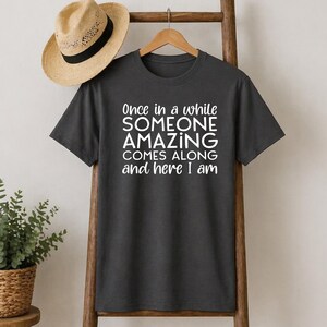 May include: Dark gray t-shirt with the text "Once in a while SOMEONE AMAZING COMES ALONG and here I am" in white. A straw hat with a black band hangs above the shirt. The shirt is displayed on a wooden rack.