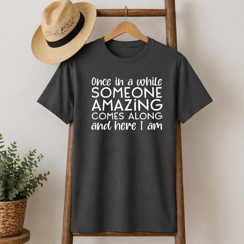 May include: Dark gray t-shirt with the text "Once in a while SOMEONE AMAZING COMES ALONG and here I am" in white. A straw hat with a black band hangs above the shirt. The shirt is displayed on a wooden rack.