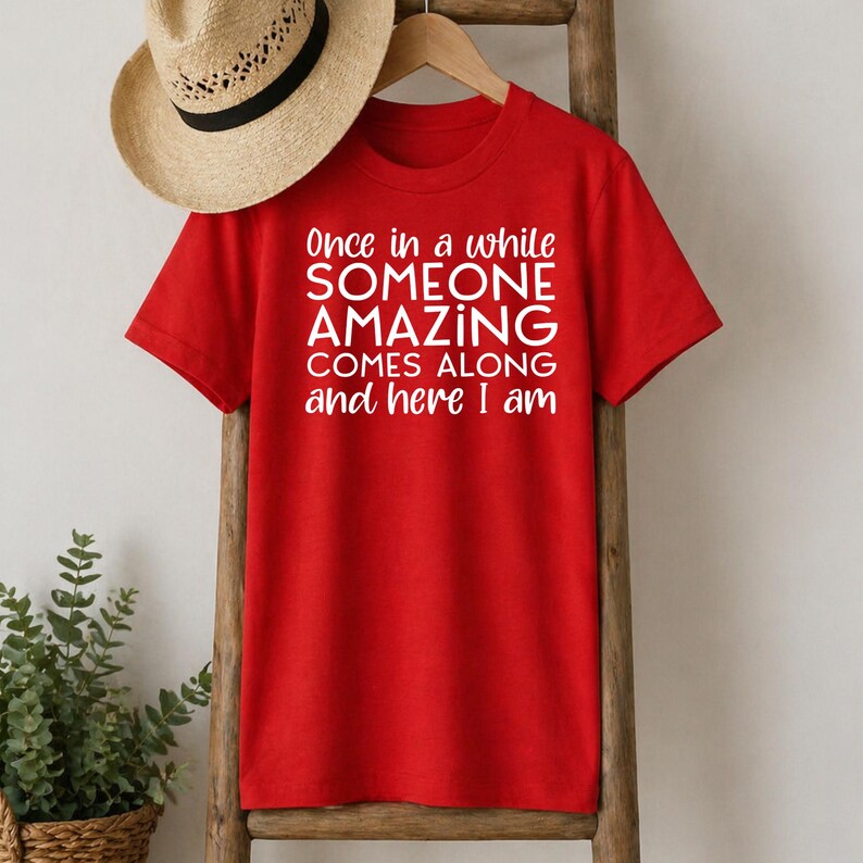 May include: A red t-shirt with the white text "Once in a while SOMEONE AMAZING COMES ALONG and here I am." A straw hat hangs above the shirt on a wooden ladder. A green plant is in a woven basket.