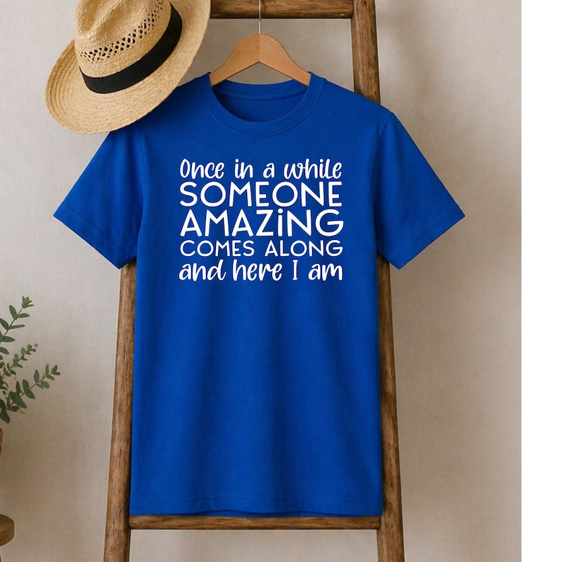 May include: A royal blue t-shirt with the text "Once in a while SOMEONE AMAZING COMES ALONG and here I am" in white. A straw hat with a black band rests on a wooden ladder. The shirt is hanging on a wooden hanger.