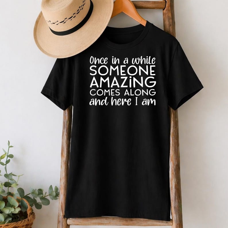 May include: Black t-shirt with the white text "Once in a while SOMEONE AMAZING COMES ALONG and here I am." A tan hat hangs above the shirt. The shirt is displayed on a wooden rack.