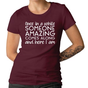 May include: A maroon t-shirt with the text "Once in a while SOMEONE AMAZING COMES ALONG and here I am" in white. The shirt is a crew neck style and has short sleeves. The image shows the shirt on a person.