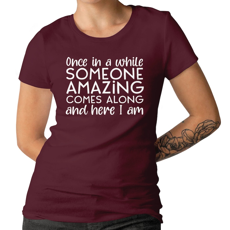 May include: A maroon t-shirt with the text "Once in a while SOMEONE AMAZING COMES ALONG and here I am" in white. The shirt is a crew neck style and has short sleeves. The image shows the shirt on a person.