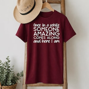 May include: A maroon t-shirt with the text "Once in a while SOMEONE AMAZING COMES ALONG and here I am" in white. A straw hat hangs above the shirt on a wooden rack.