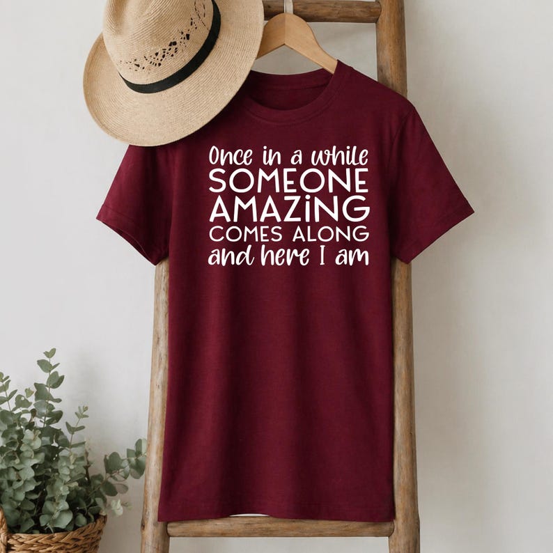 May include: A maroon t-shirt with the text "Once in a while SOMEONE AMAZING COMES ALONG and here I am" in white. A straw hat hangs above the shirt on a wooden rack.