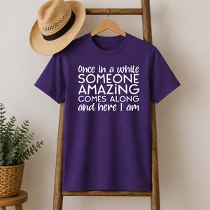 May include: A purple t-shirt with the text "Once in a while SOMEONE AMAZING COMES ALONG and here I am" in white. A straw hat with a black band hangs above the shirt.