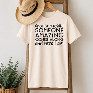 May include: A cream-colored t-shirt with the text "Once in a while SOMEONE AMAZING COMES ALONG and here I am" in black. A straw hat with a black band hangs above the shirt.