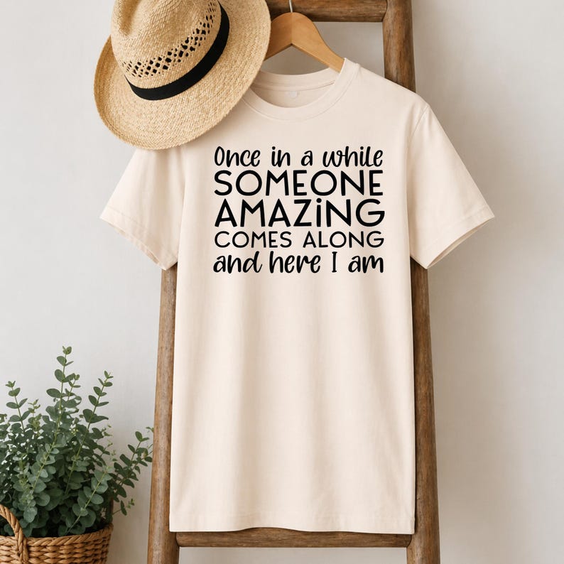 May include: A cream-colored t-shirt with the text "Once in a while SOMEONE AMAZING COMES ALONG and here I am" in black. A straw hat with a black band hangs above the shirt.