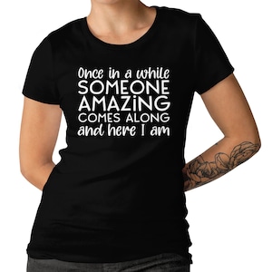 May include: Black t-shirt with the white text: "Once in a while SOMEONE AMAZING COMES ALONG and here I am." The shirt has a crew neck and short sleeves. The person wearing the shirt has a tattoo on their arm.