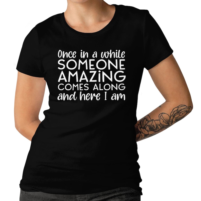May include: Black t-shirt with the white text: "Once in a while SOMEONE AMAZING COMES ALONG and here I am." The shirt has a crew neck and short sleeves. The person wearing the shirt has a tattoo on their arm.