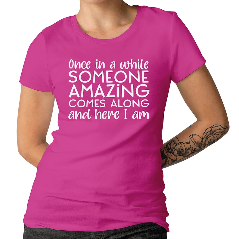 May include: A magenta t-shirt with the text "Once in a while SOMEONE AMAZING COMES ALONG and here I am" in white. The shirt has short sleeves and a crew neck.