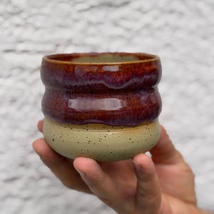 May include: A handmade ceramic tumbler with a unique design. The top section is a glossy, deep burgundy color, while the bottom is a speckled beige. The interior has a green hue. The tumbler is held in a hand, showcasing its size and shape.
