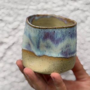 Puede incluir: Vaso de cerámica hecho a mano con un diseño único. La mitad inferior es de color beige claro, mientras que la mitad superior presenta una mezcla de tonos azules, morados y blancos. El borde tiene un aspecto natural y sin esmaltar.