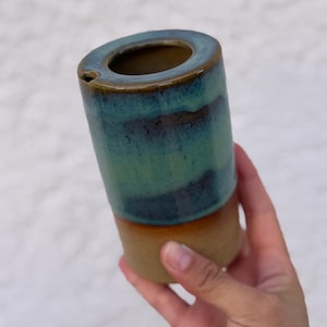 Op de afbeelding: Een cilindervormig keramieken vat met een tweekleurig glazuur. Het bovenste gedeelte heeft een glanzende, gevlekte afwerking in tinten blauw, groenblauw en donkerbruin, terwijl de onderkant een matte, ongeglazuurde afwerking van beige klei heeft.