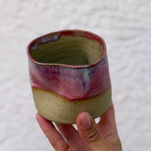 May include: A handmade ceramic cup with a speckled beige base and a vibrant pink and purple glazed rim. The vessel features an organic, slightly irregular shape with a smooth texture on the upper portion and a matte finish on the lower section.