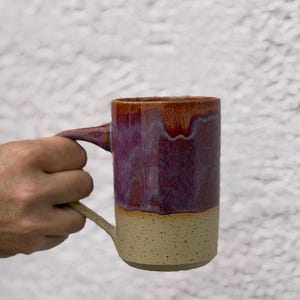 May include: A handmade ceramic mug with a large handle. The top half of the mug is a deep purple with reddish-brown accents, while the bottom half is a speckled beige. The mug is held by a person.