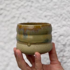 Könnte beinhalten: Eine handgefertigte Keramiktasse mit einem gesprenkelten, hellbeigen Boden und einem geschichteten, strukturierten oberen Bereich. Der obere Bereich weist Gelb-, Grün- und Brauntöne mit glänzendem Finish auf. Die Tasse wird in einer Hand gehalten.