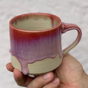 Könnte beinhalten: Eine handgefertigte Keramiktasse mit einer einzigartigen Glasur. Die Tasse weist einen Farbverlauf auf, der von einem tiefen Rot am Rand zu einem hellen Lila und Beige am Boden übergeht. Die Glasur hat einen Tropfeffekt. Die Tasse wird in einer Hand gehalten.