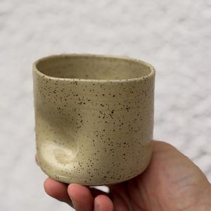 Puede incluir: Una taza de cerámica beige moteada, con una hendidura curva única en un lado. La taza cilíndrica está salpicada de pequeñas manchas oscuras. La taza se sostiene en una mano. El diseño es táctil.