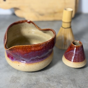 Può includere: Una ciotola per matcha in ceramica fatta a mano, un porta frusta e una frusta in bambù. La ciotola ha una forma unica con una glassa rosso-viola su una base beige. Il porta frusta è a forma di cono.