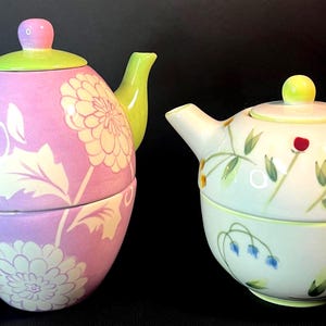 May include: Two ceramic teacup and teapot sets. One set is lavender with white floral designs and green accents. The other is white with colorful floral designs and green accents. Both sets are stacked.