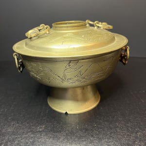 May include: A vintage brass hot pot with a lid and two handles. The pot has an engraved design of birds and mountains. The pot sits on a round base.