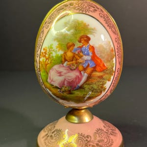 May include: A porcelain egg-shaped decorative object with a gold-trimmed base and a painted scene of a couple in a garden. The egg is on a gold stand with a pink base.