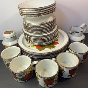 May include: A vintage ceramic dinnerware set featuring white plates, bowls, and cups with a brown rim. The plates and cups are decorated with a strawberry design. Includes a creamer and a sugar bowl.