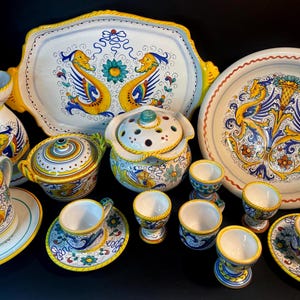 May include: A collection of hand-painted ceramic tableware featuring a vibrant design with yellow, blue, and white accents. The set includes a serving tray, plates, cups, saucers, and egg cups. The decorative style is reminiscent of Italian pottery.