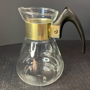 May include: A clear glass coffee pot with a gold band around the top and a black plastic handle. The pot has a round body and a wide mouth. The text "HEAT RESISTANT" is visible on the bottom of the pot.