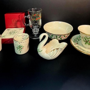 May include: A collection of holiday-themed tableware, including a teacup and saucer, bowl, swan-shaped dish, and two candle holders. The pieces are cream-colored with gold trim and holly berry designs. A glass mug and a red box are also present.