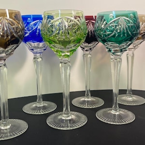 May include: A collection of eight colorful crystal wine glasses. Each glass features a different colored bowl, including red, amber, blue, green, and teal. The glasses have long, clear stems and circular bases, with intricate cut patterns.