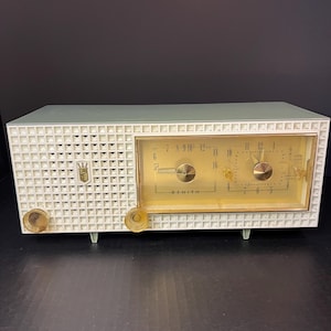 May include: A vintage Zenith radio with a white and green plastic case. The radio has a gold-colored dial with black numbers and a white needle. The radio has a white plastic grille with a diamond pattern. The radio has a gold-colored knob on the right side.