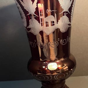 May include: A burgundy-colored glass vase with white etched floral and architectural designs. The vase has a bulbous base and a flared top, with a decorative pattern around the rim and base. A sticker is visible near the base.